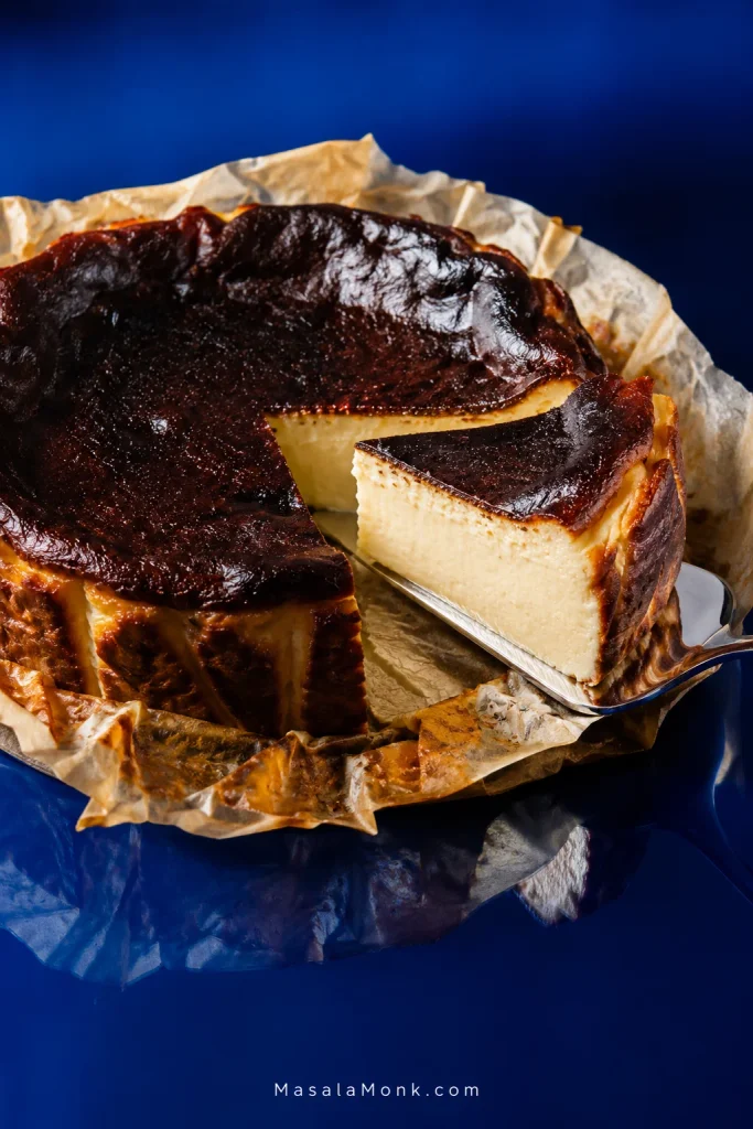 Basque cheesecake slice lifted on a silver server, showing a pale creamy center beneath a dark caramelized top.