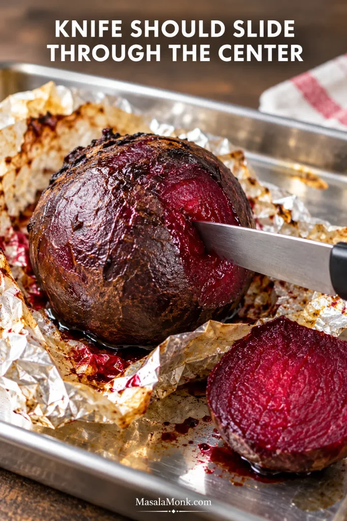 Thin knife inserted through the thick center of a roasted red beet on a metal tray, exposing moist, fully cooked ruby-red flesh.
