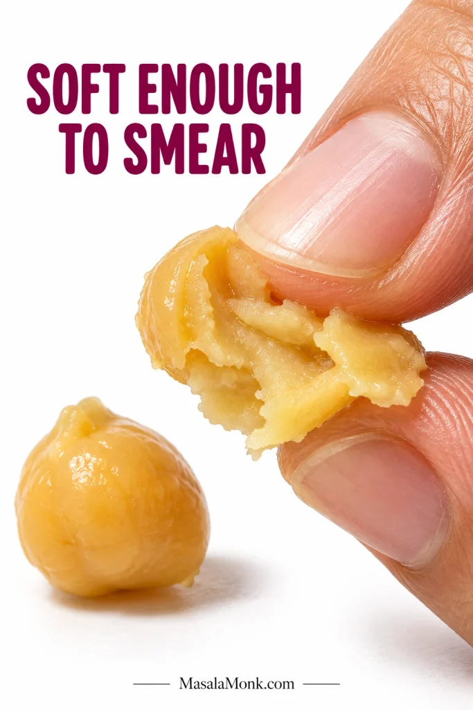 Fingertips pressing a cooked chickpea into a soft smear against a white background, demonstrating a properly softened chickpea.
