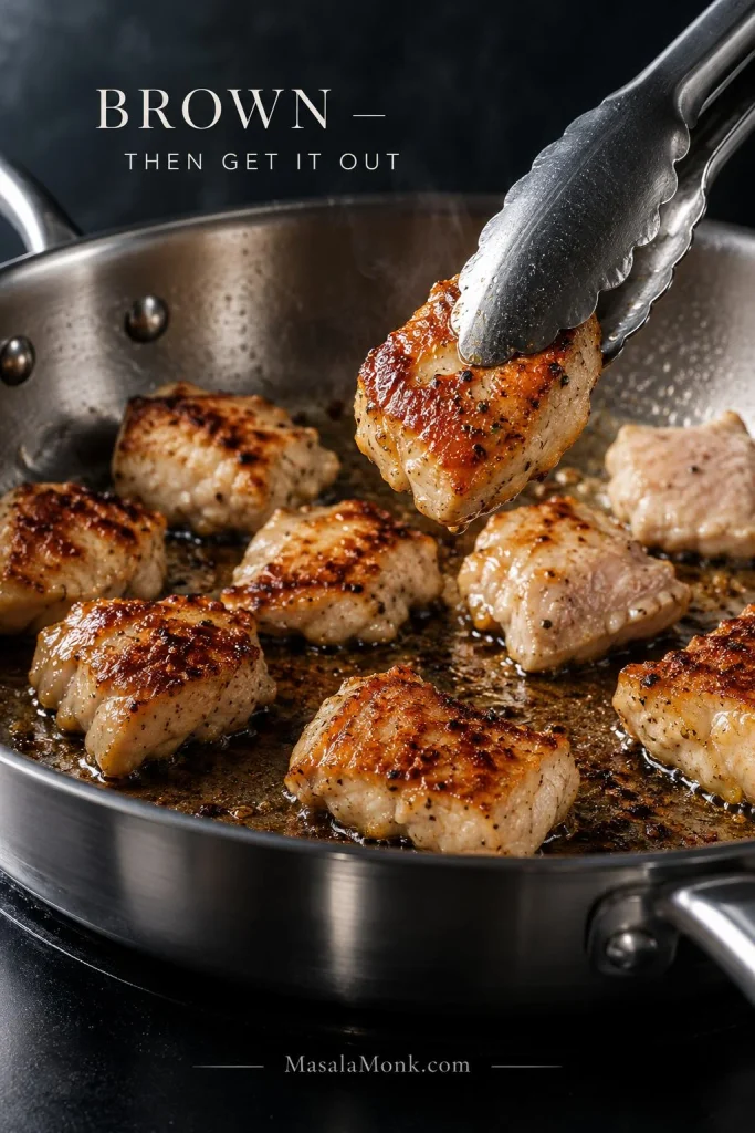 Metal tongs lift a piece of chicken with deep golden-brown searing from a skillet containing browned and paler chicken pieces.