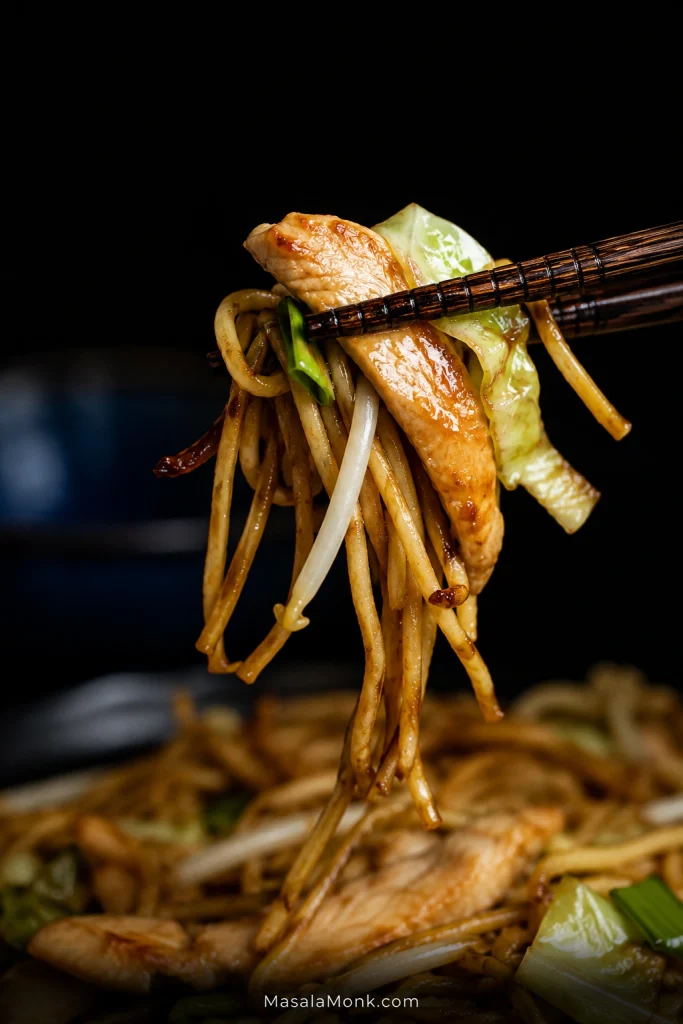 Close-up chopsticks lifting glossy chow mein noodles with a thin browned chicken strip, cabbage, bean sprout and scallion against a dark background.