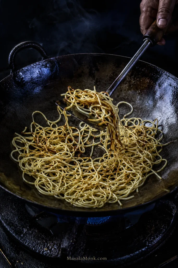 Medium-thin yellow chow mein noodles spread across a dark wok with exposed metal and scattered golden-brown toasted patches, without chicken, vegetables or sauce.