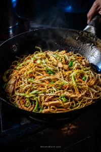 Chicken chow mein in a dark carbon-steel wok with thin chicken strips, medium-thin noodles, green cabbage, bean sprouts, carrots and scallions, with glossy strands and scattered browned edges.
