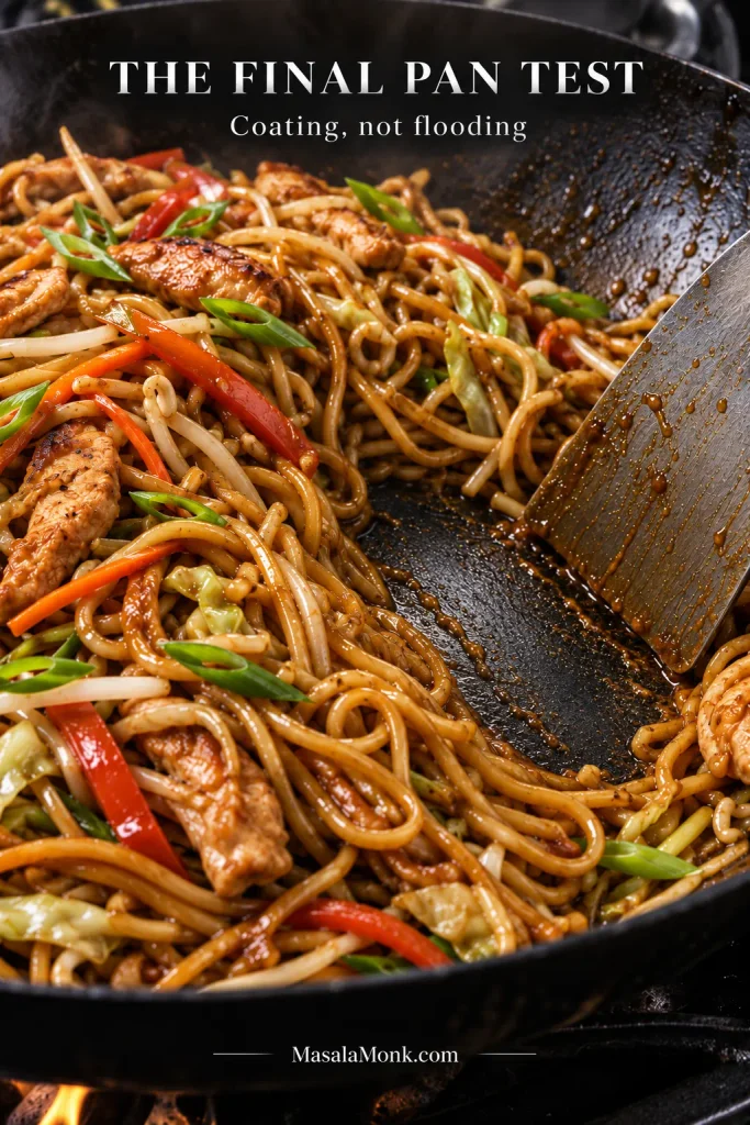 Close-up of chicken lo mein in a dark wok with a metal spatula clearing a channel through the noodles and exposing a thin layer of glossy sauce on the pan.