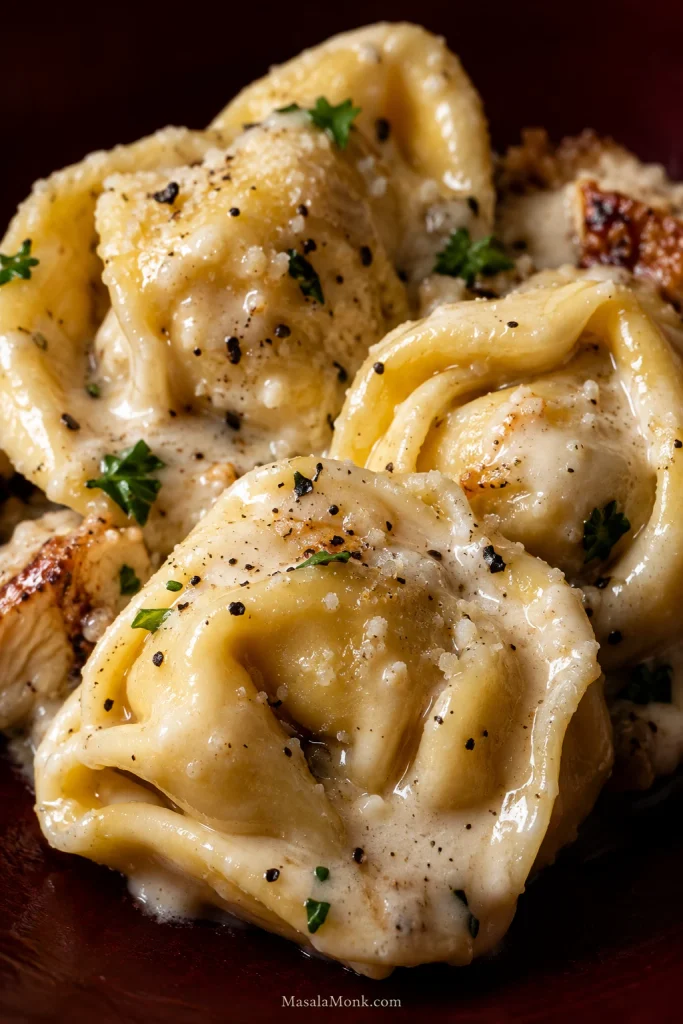 Close-up of cheese tortellini and browned chicken lightly coated in glossy Alfredo sauce, with the pasta folds still visible.
