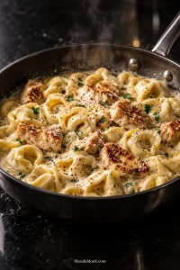 A dark skillet filled with cheese tortellini, browned chicken pieces and glossy Alfredo sauce, finished with black pepper, finely grated Parmesan and a small amount of parsley.