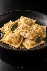 Plate of pale golden homemade cheese ravioli coated lightly in brown butter and Parmesan, with one raviolo cut open to show creamy ricotta filling.