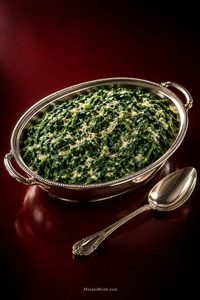 Creamed spinach with visible leafy greens coated in a smooth cream sauce, served in an oval silver dish on a deep red background.