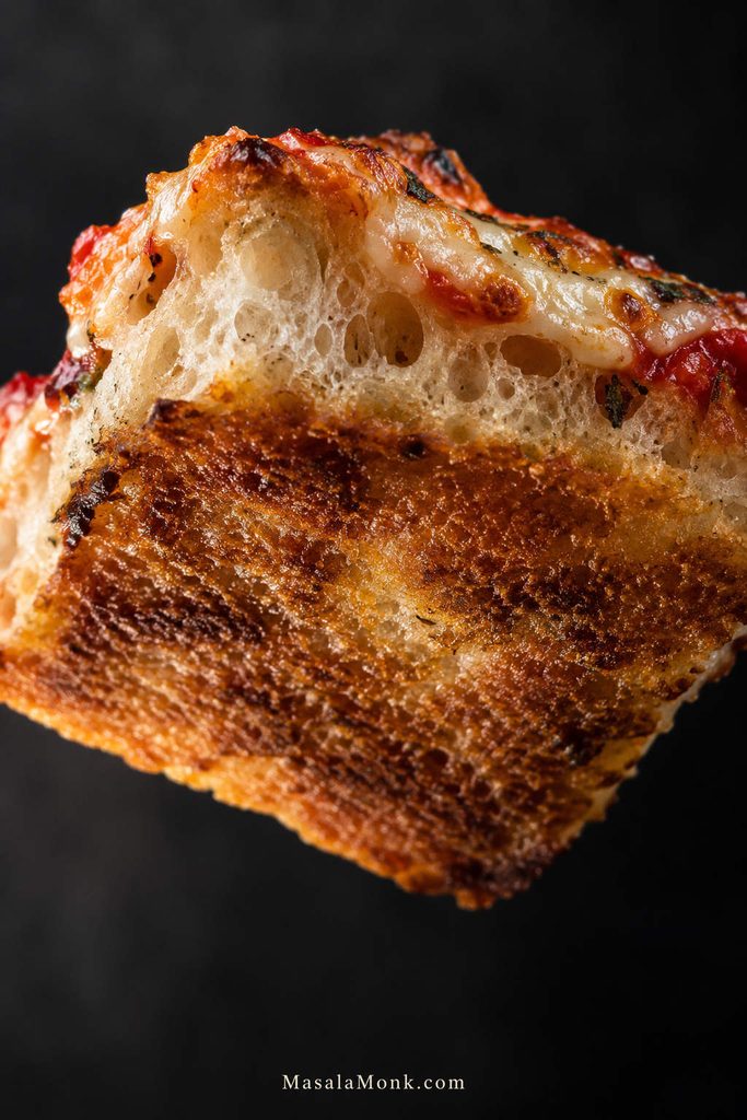 Focaccia pizza slice tilted upward to show a deeply browned underside, airy crumb, tomato sauce, and melted cheese.