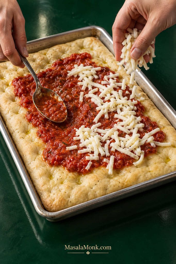 Thick tomato sauce and shredded mozzarella being added in a restrained layer to par-baked focaccia in a metal sheet pan.