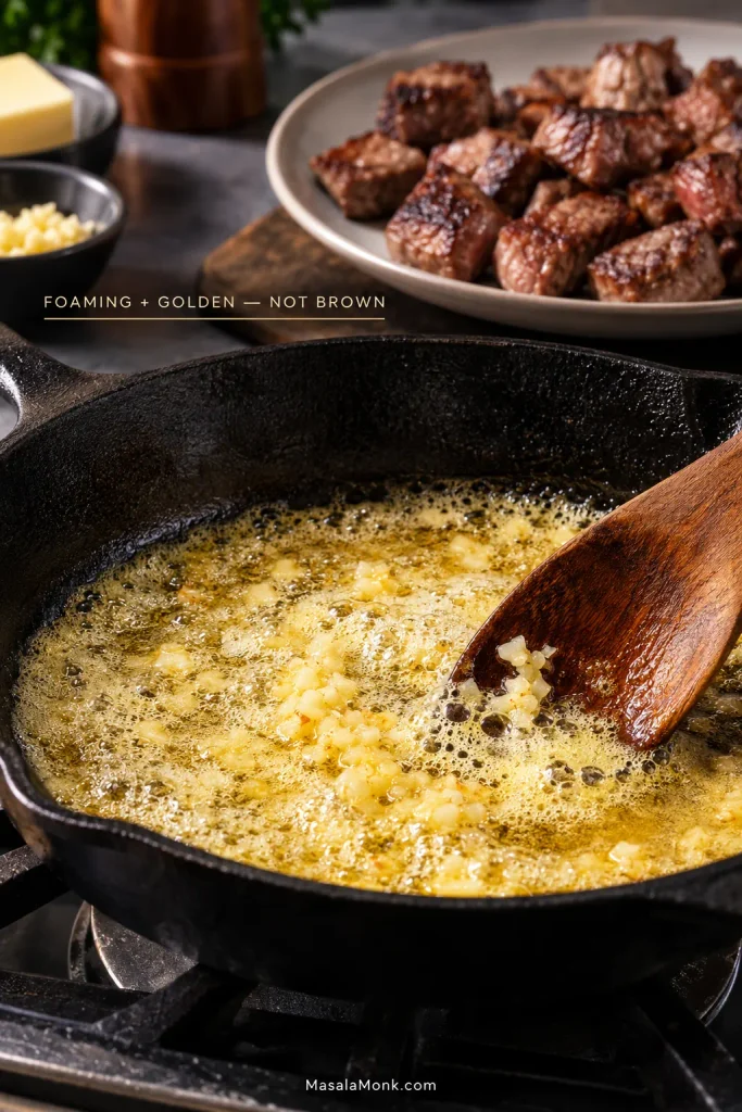Golden foaming butter and pale minced garlic in a cast-iron skillet, with browned steak bites resting on a plate behind it.