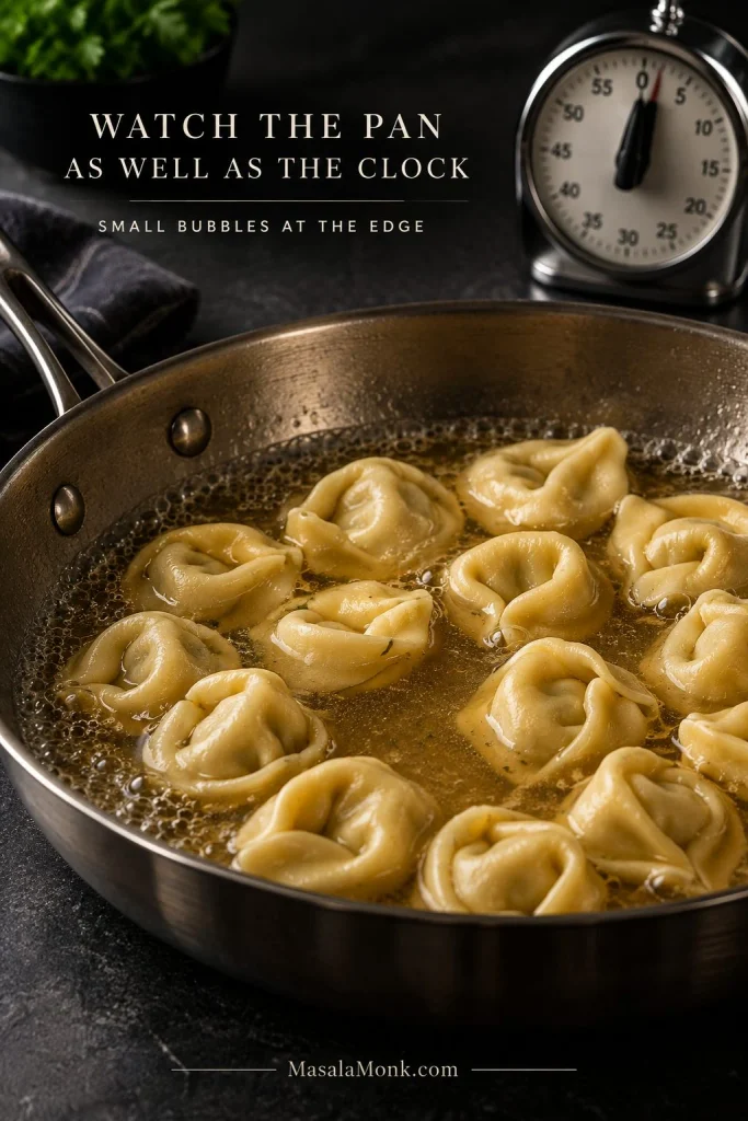 Cheese tortellini gently simmering in shallow broth with small bubbles around the skillet edge; text reads “Watch the Pan as Well as the Clock.”