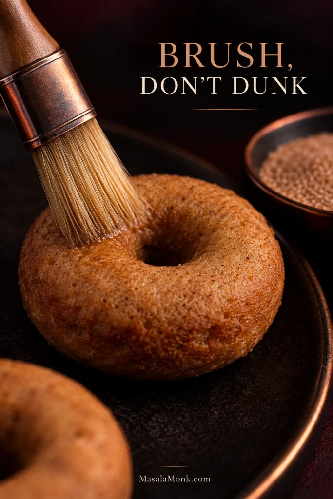 Pastry brush applying a thin layer of butter to a plain baked donut, with a bowl of cinnamon sugar nearby.