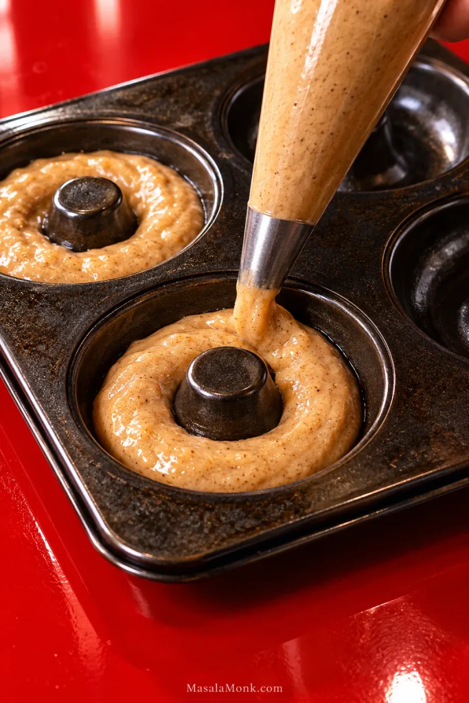 Piping bag filling a dark metal donut pan with thick speckled batter on a glossy red surface.