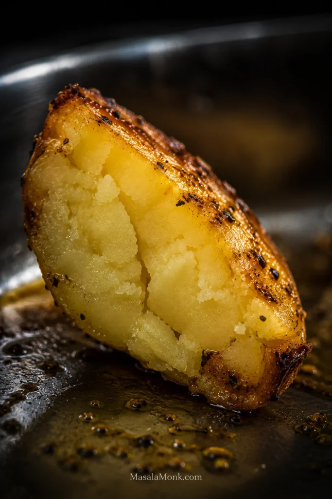 Close-up of a broken roasted potato wedge showing a creamy pale-yellow center, browned outer edge, oregano flecks and a light sheen of lemon-garlic pan juices.