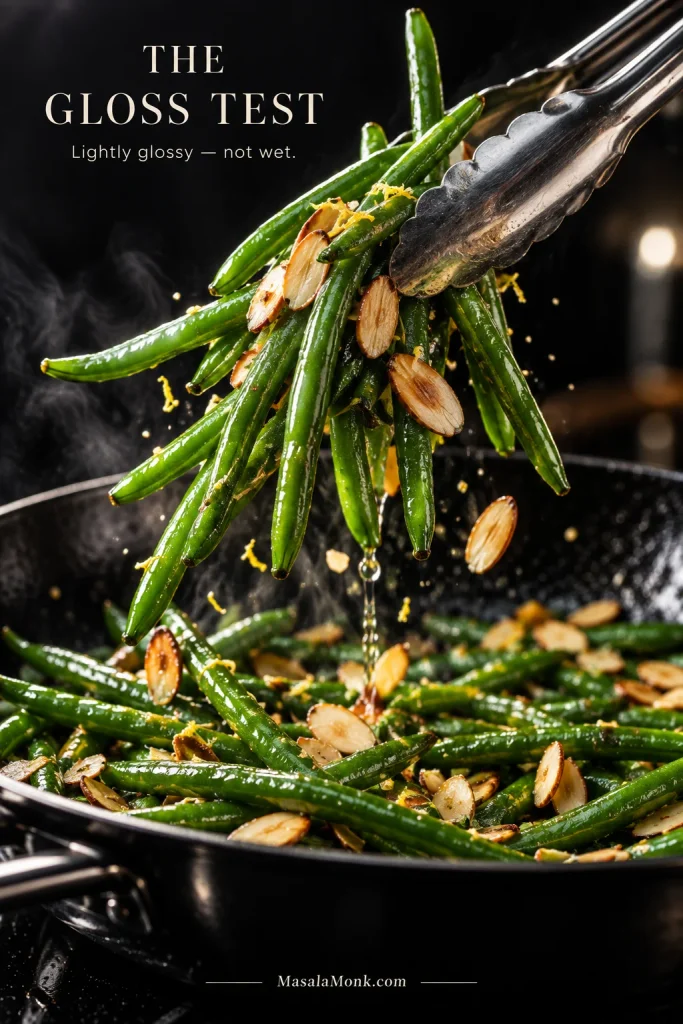 Metal tongs lifting glossy green beans and toasted almonds from a dark skillet with no visible pool of butter or sauce beneath them.
