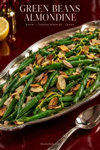 Silver platter of glossy green beans almondine topped with toasted sliced almonds, translucent shallot and fine lemon zest on a deep oxblood table.