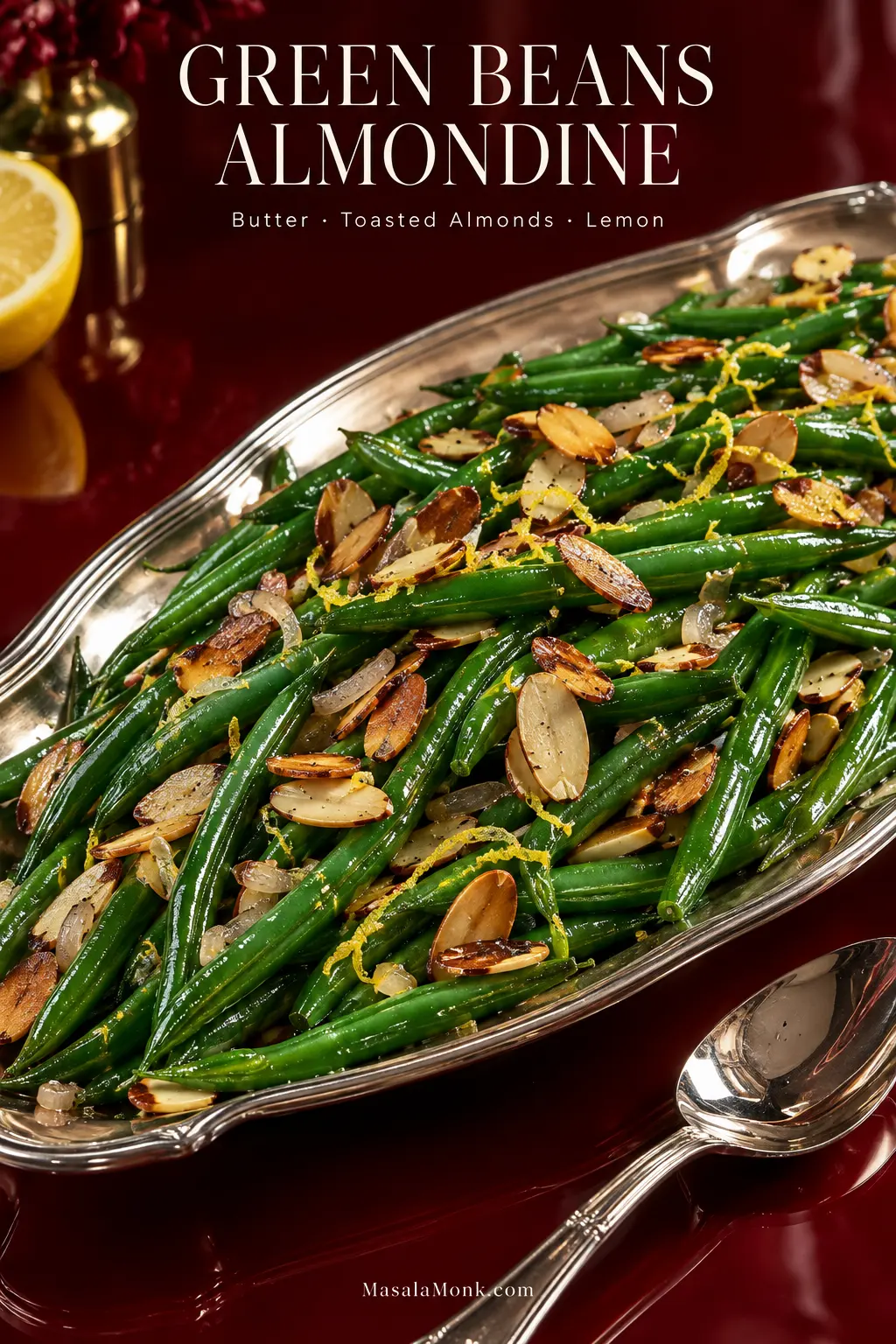 Silver platter of glossy green beans almondine topped with toasted sliced almonds, translucent shallot and fine lemon zest on a deep oxblood table.