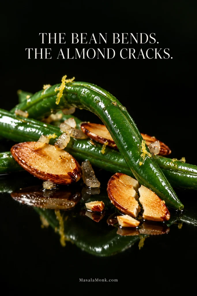 Macro view of curved glossy green beans with broken toasted almond pieces on a black reflective surface beneath the words “The bean bends. The almond cracks.”