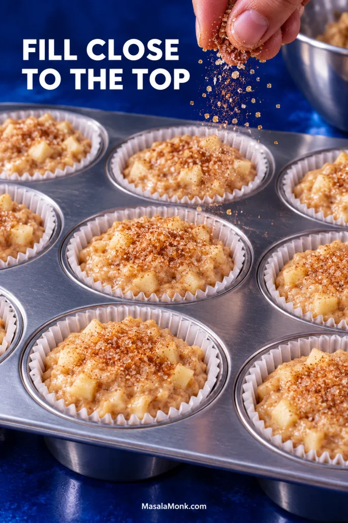 Apple muffin batter portioned close to the rims of white paper liners in a muffin pan while cinnamon sugar is added on top.