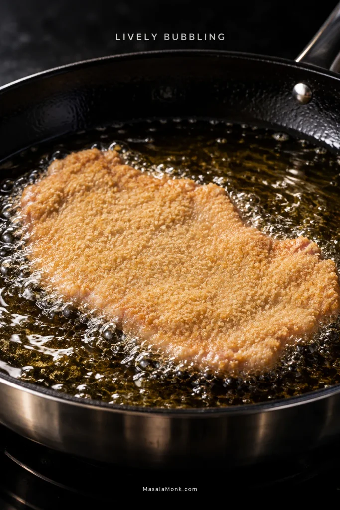 Thin breaded pork schnitzel frying in shallow hot oil with active bubbles forming around its edges in a skillet.