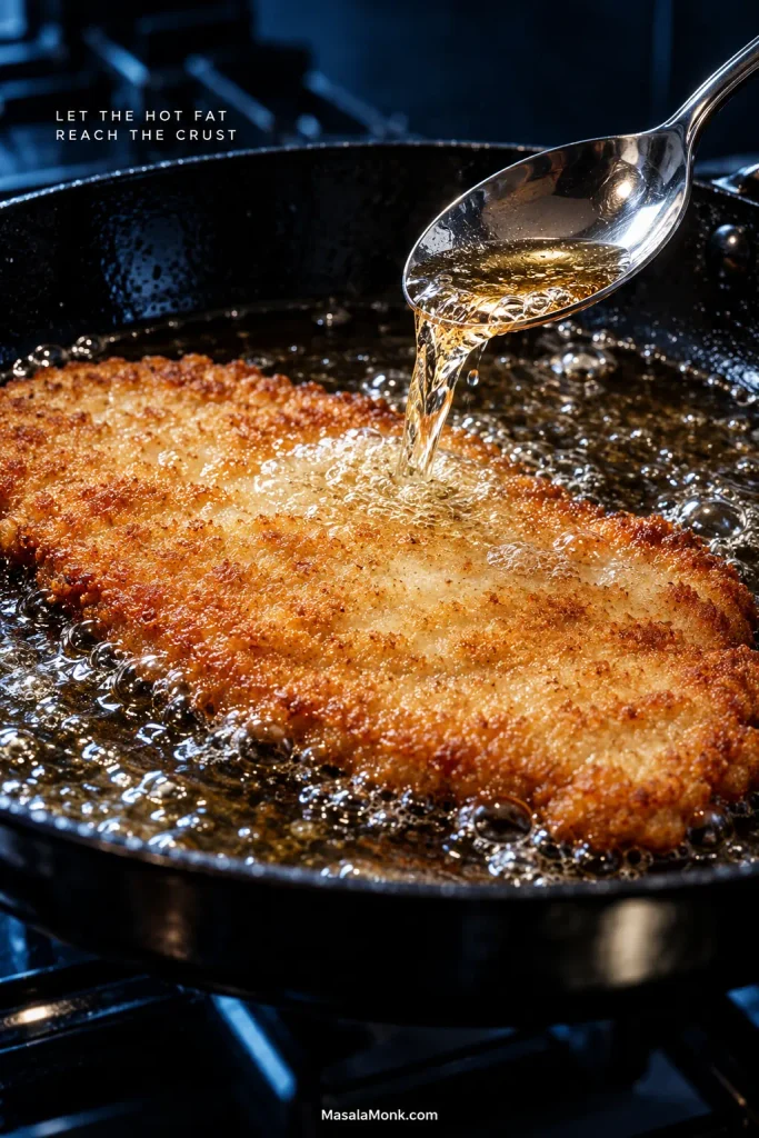 Metal spoon lifting hot frying oil from the skillet and pouring it over a pale area of a thin pork schnitzel as it shallow-fries.