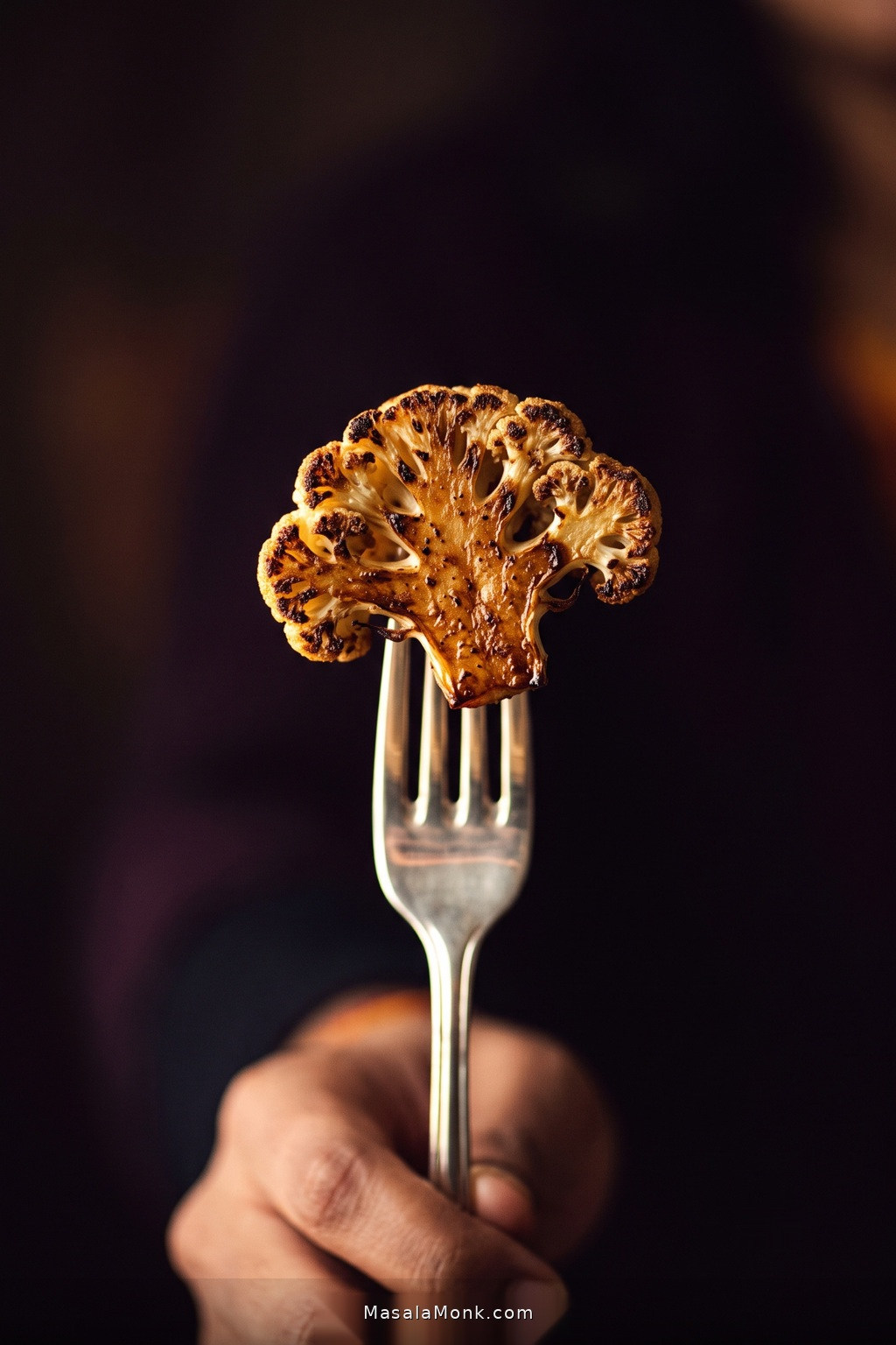 A silver fork lifts one deeply roasted cauliflower piece toward the viewer, showing dark browned edges and a pale tender interior against a deep plum background.
