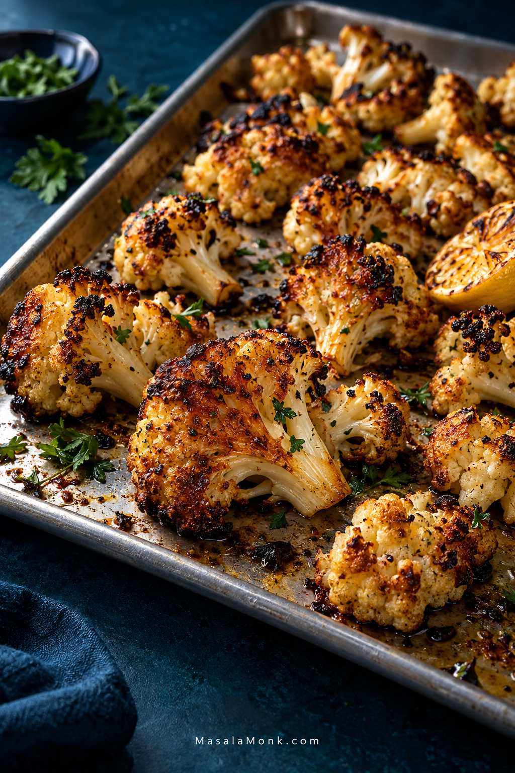 A brushed-metal sheet pan filled with irregular roasted cauliflower florets, showing deep golden-brown edges, darker roasted tips, tender stems, fresh green herbs, and a small lemon garnish against a deep teal background.