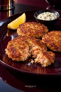 Three broad, thin golden-brown salmon patties on a dark plate; one is torn open to show moist pink salmon flakes, with lemon and tartar sauce behind.