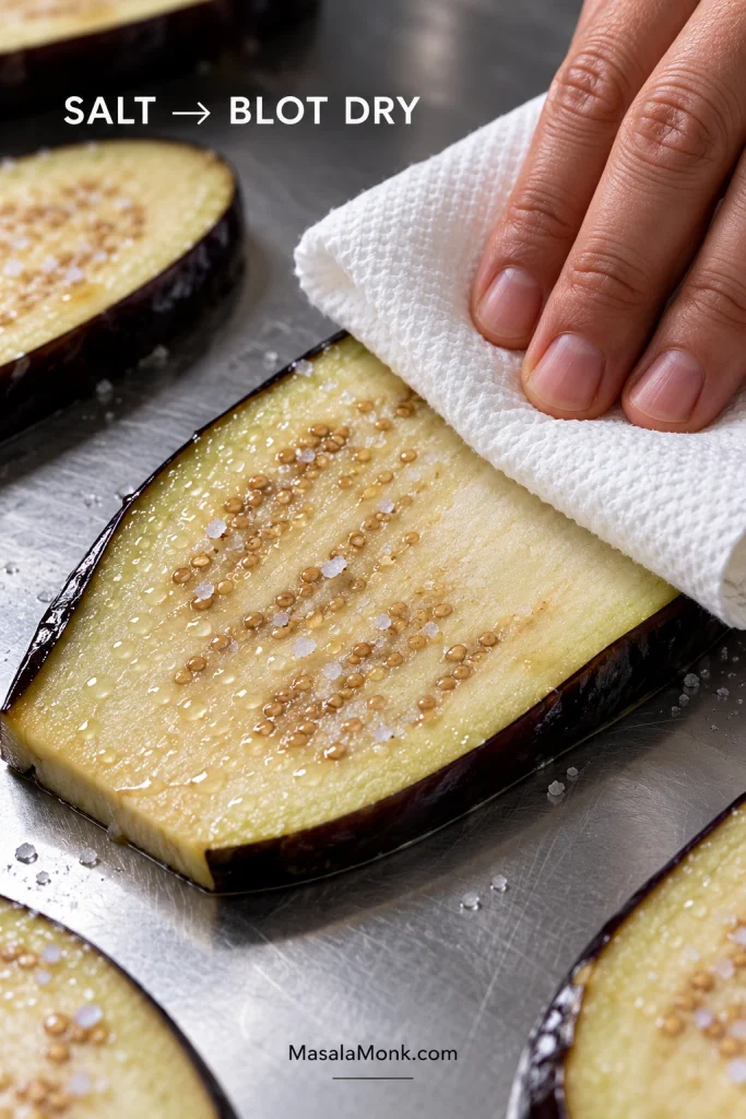 Hand blotting moisture from a salted lengthwise eggplant slice with a white towel before roasting.