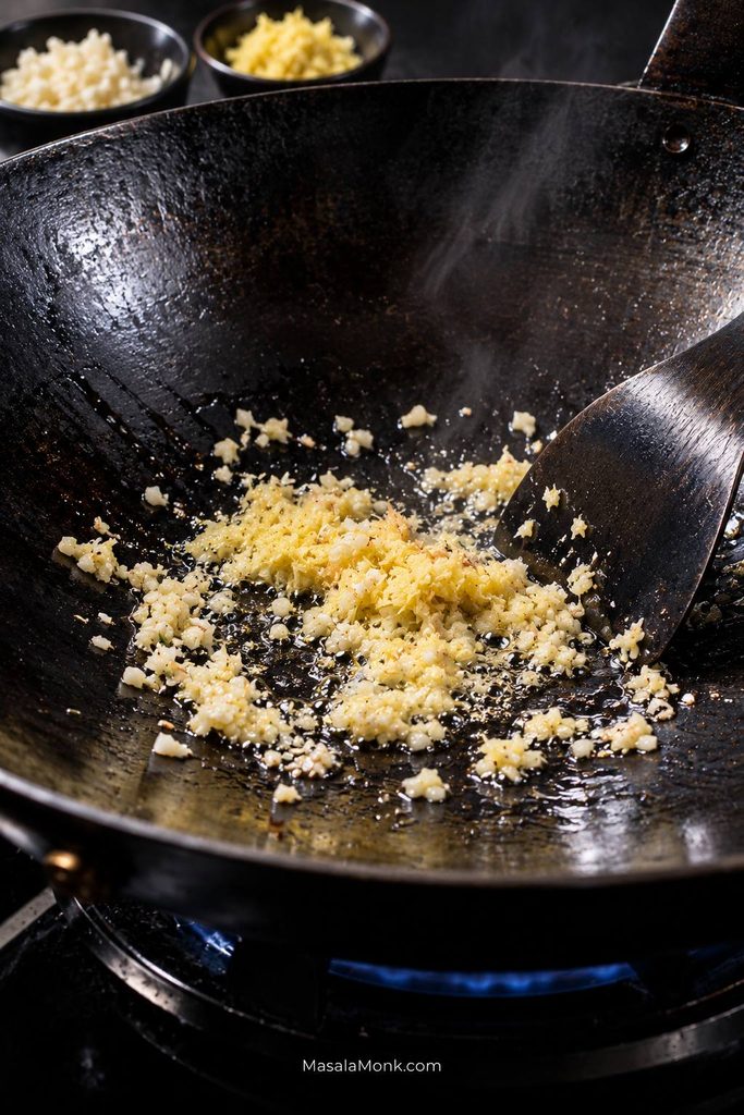 Finely minced garlic and grated ginger sizzling briefly in a thin film of oil in a dark wok over a blue gas flame.
