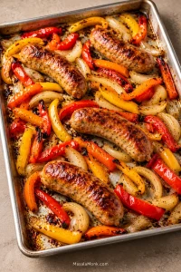 Roasted Italian sausage links with red, orange and yellow bell pepper strips and golden onion spread across a rimmed aluminum sheet pan with visible areas of bare metal.