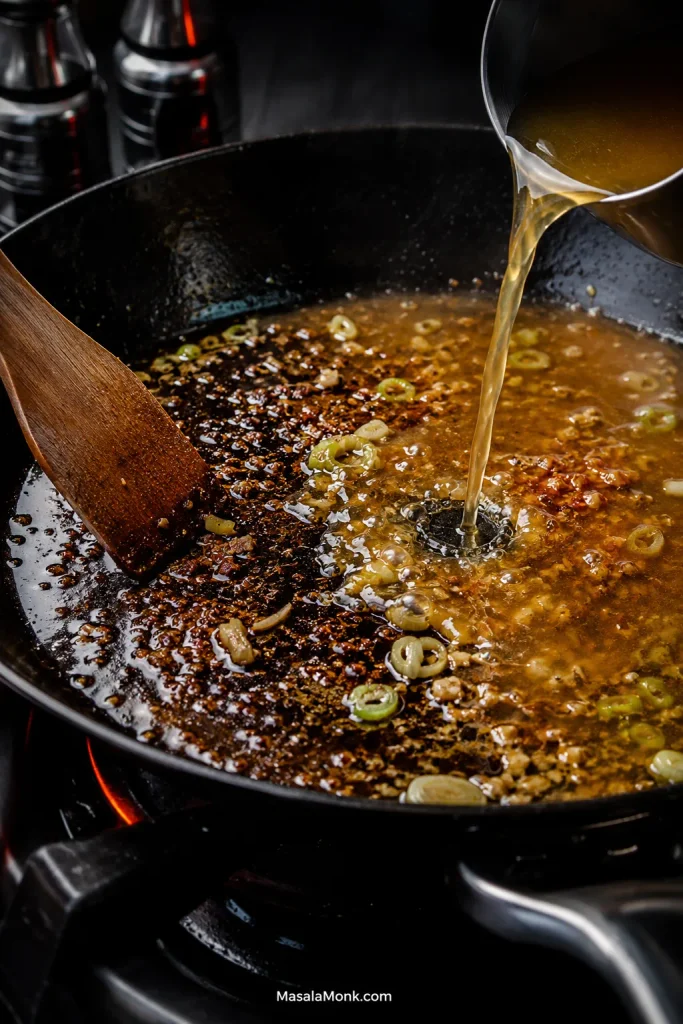 Broth poured into a skillet with browned fond, garlic and scallions while a spatula scrapes the pan into glossy gravy.