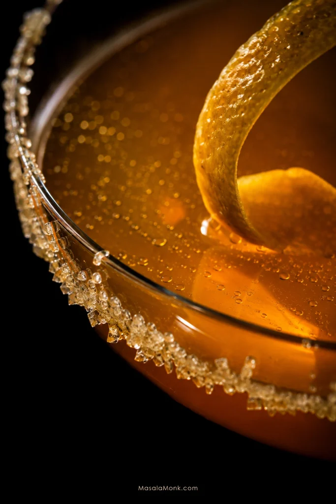 Macro view of a Sidecar with amber liquid, a partial sugar rim, lemon twist, citrus-oil droplets and tiny post-shake bubbles.