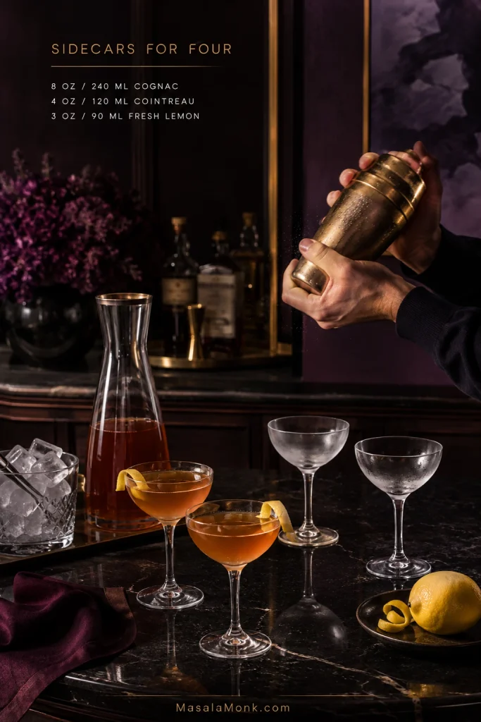 Hands shaking a cocktail beside a prebatch carafe, two finished Sidecars and two chilled empty coupes for a four-serving batch.