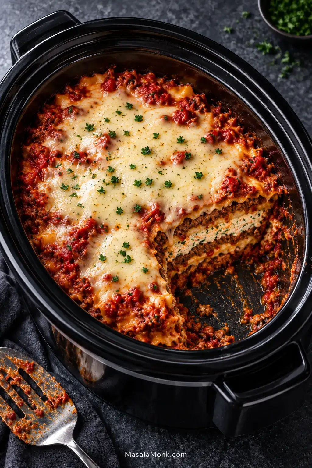 Finished lasagna in a black oval slow cooker with one serving removed, revealing pasta, red meat sauce, creamy ricotta and melted mozzarella.