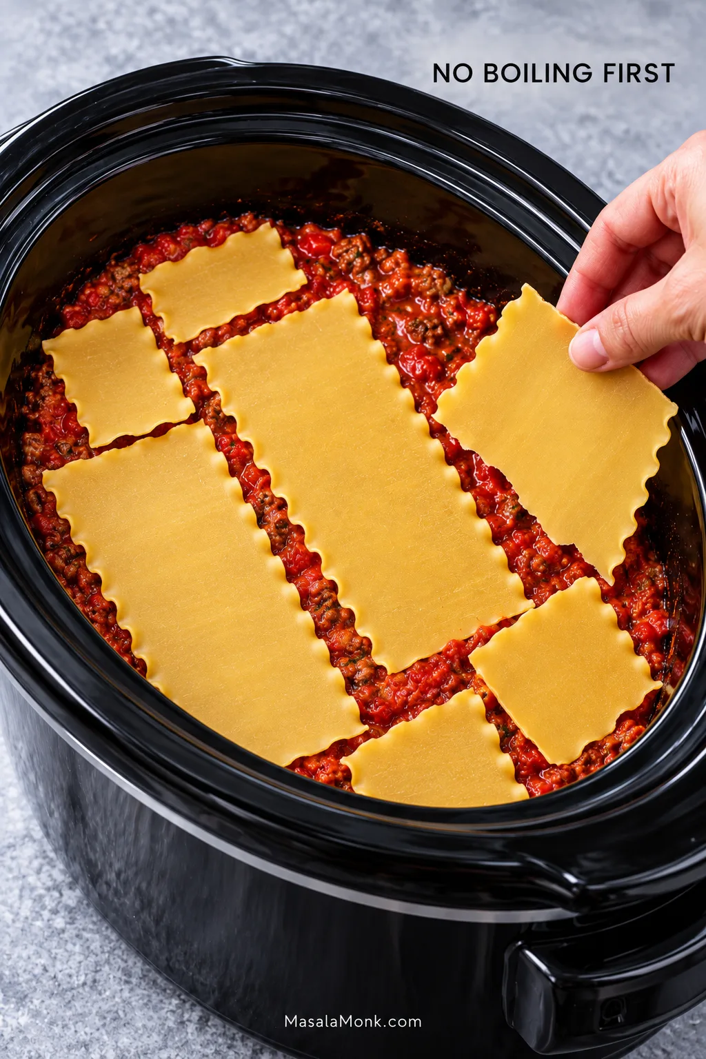 Dry lasagna noodles being placed over meat sauce in a black oval slow cooker, with several sheets broken into smaller pieces to fit the curved crock.