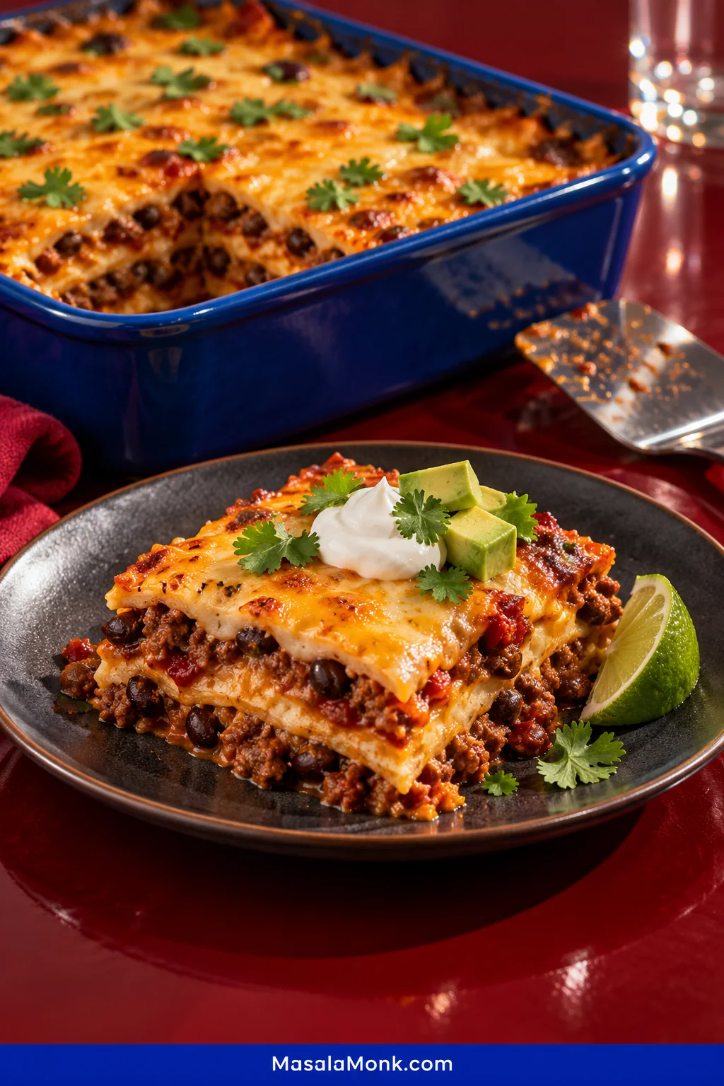 A square serving of layered taco casserole with flour tortillas, seasoned ground beef, black beans, salsa and melted cheese, topped with sour cream, avocado and cilantro, with the remaining casserole in a blue baking dish behind it.