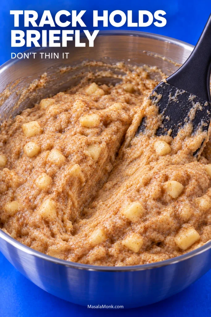 Thick apple muffin batter with visible apple pieces in a metal bowl, showing ridges and a channel made by a spatula.
