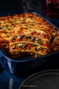 Finished vegetable lasagna in a deep blue baking dish with a lifted slice showing pasta, ricotta, spinach, zucchini, mushrooms, peppers and tomato sauce.