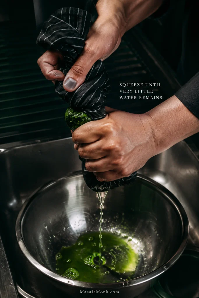 Hands twisting cooked spinach in a dark towel over a stainless-steel bowl to squeeze out excess water.