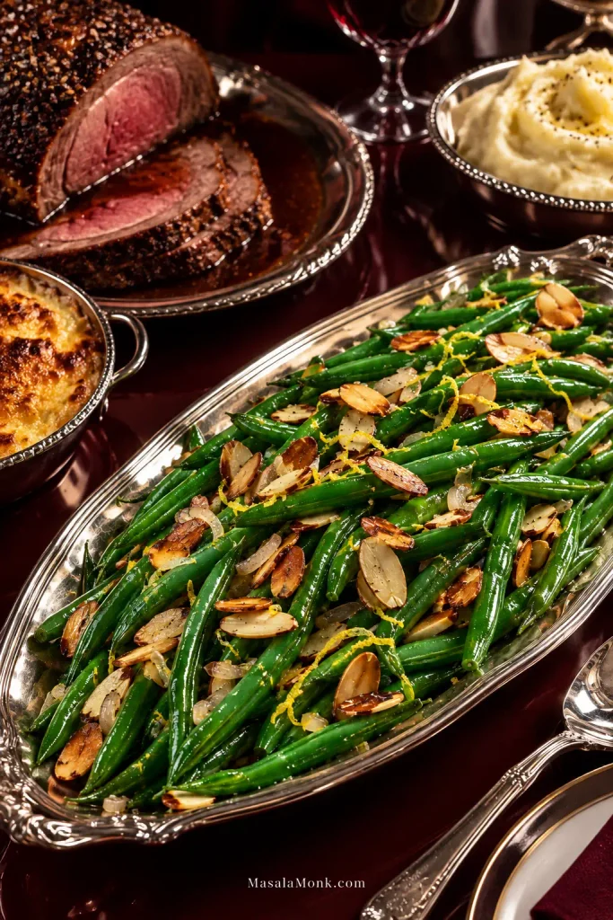 Formal dinner table with a large silver platter of green beans almondine in the foreground, alongside sliced roast beef, mashed potatoes and a baked side dish.