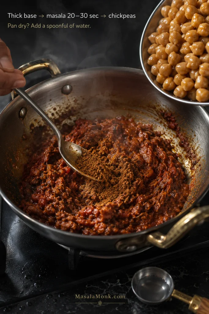 Spoon stirring chana masala powder into a thick cooked onion-tomato base, with chickpeas ready to be added and a small measure of water nearby.