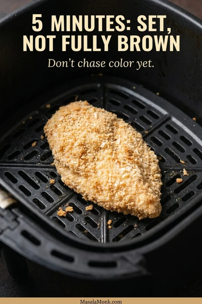 Partially cooked breaded chicken breast in an air fryer with set pale crumbs and scattered early golden browning.