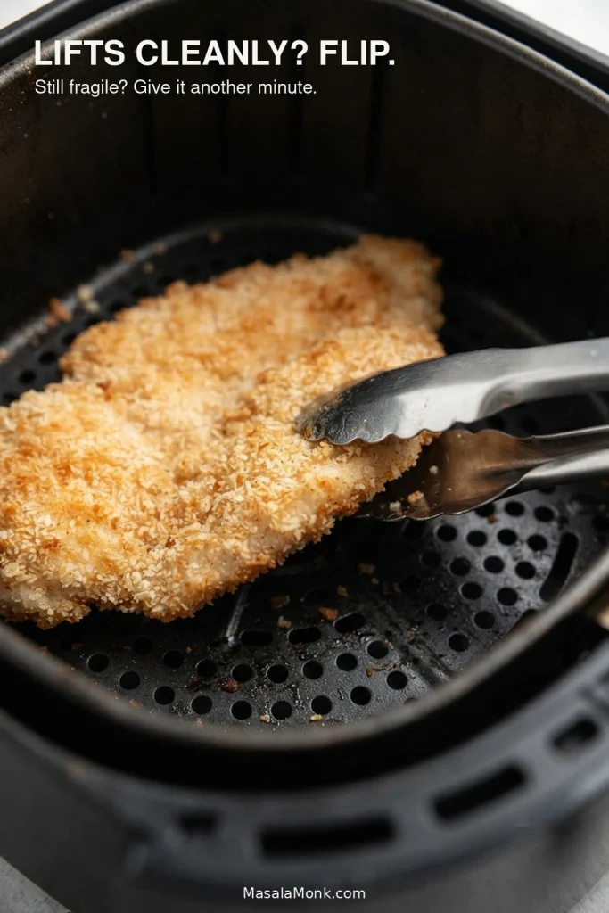 Stainless-steel tongs lifting one edge of a breaded chicken breast to check whether the crust releases from the air fryer basket.