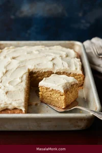 Rectangular banana sheet cake topped with cream cheese frosting in a metal pan, with one square removed to reveal the golden crumb.