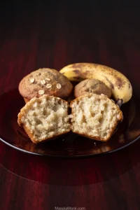 Two banana oatmeal muffins split open on a dark plate, showing a pale tender crumb, with two whole muffins and a spotted ripe banana behind them.