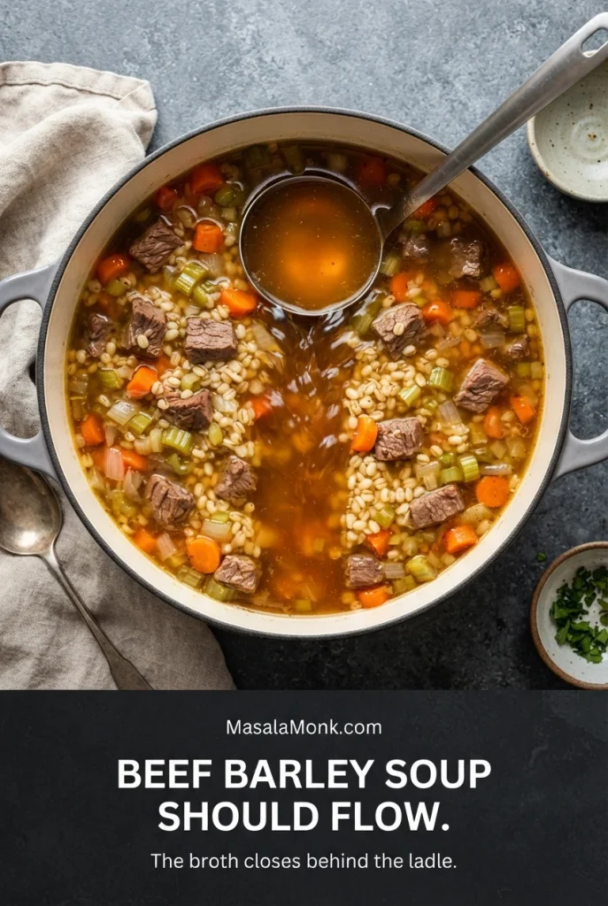 Overhead pot of beef barley soup with a ladle raised above the surface, showing flowing brown broth between beef, barley and vegetables.