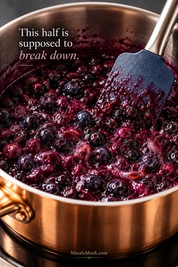 Blueberries cooking in a saucepan, with split and collapsed berries releasing dark purple juice while some berries remain recognizable.