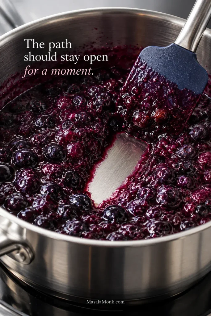 Spatula drawn through thickened blueberry filling in a saucepan, leaving a temporary clear path across the bottom.